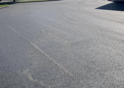Freshly paved parking lot with several cars and a tree line in the background, under a sunny sky.
