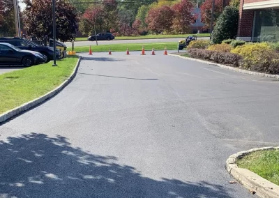 A row of orange traffic cones blocks a paved driveway near a commercial building with trees in the background.