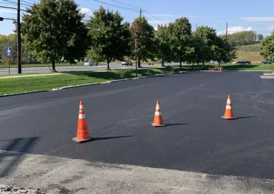 Three orange traffic cones block off a newly paved asphalt area near green trees and a road.