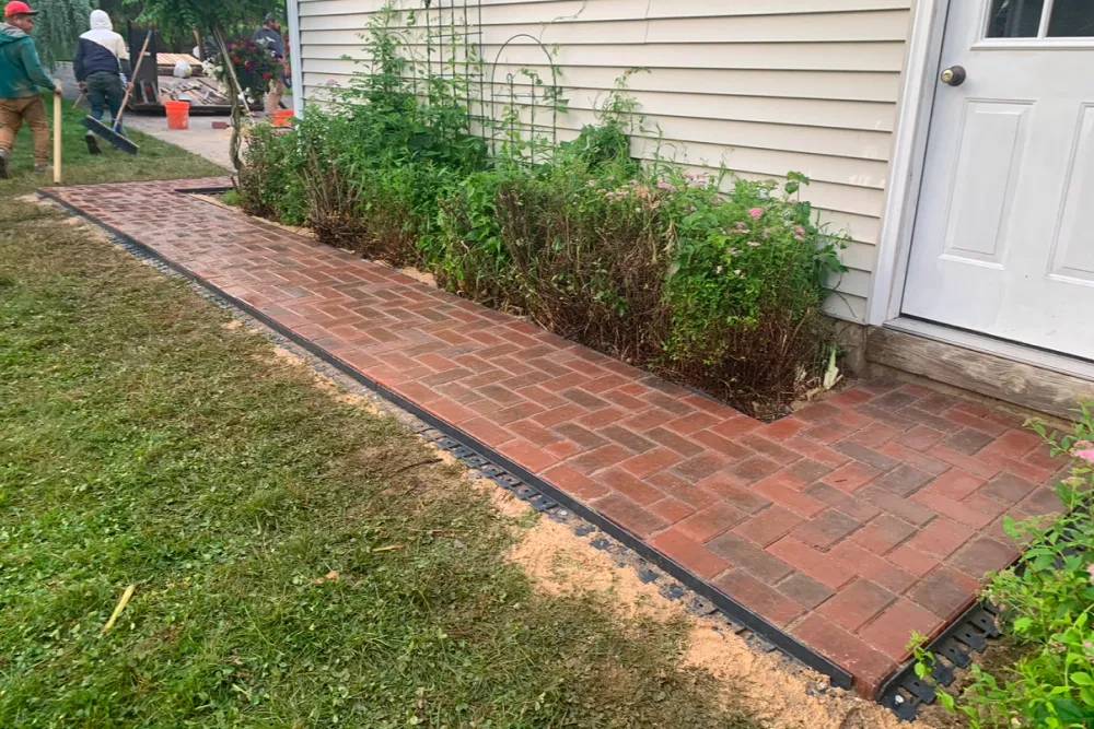 Red brick walkway beside a house, bordered by plants, with workers visible in the background.