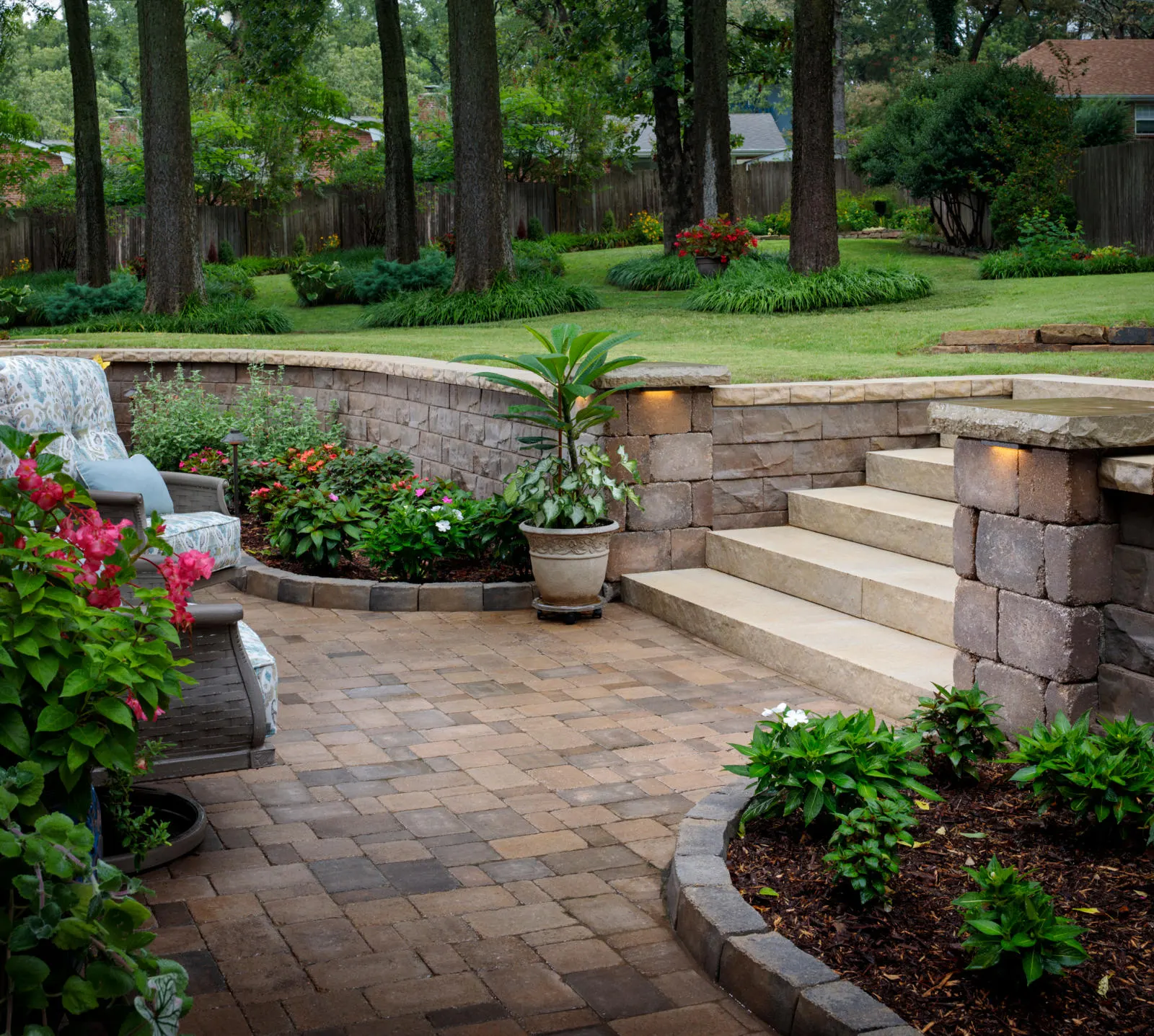 Brick patio with potted plants, stone steps, and garden seating, overlooking a green, landscaped backyard.