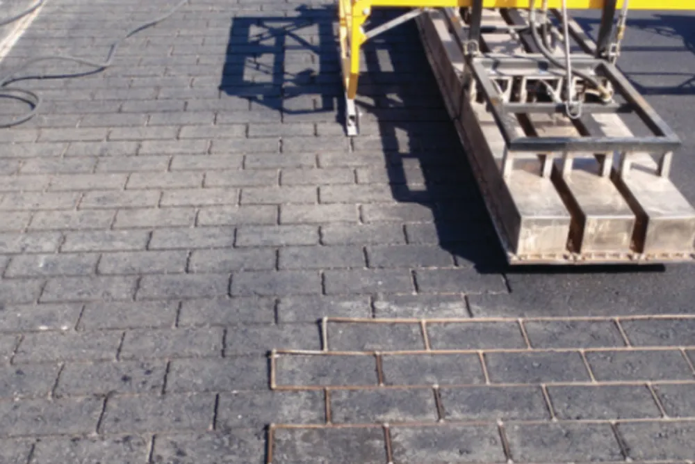 A machine is laying rectangular concrete pavers on a surface, creating a patterned pavement.