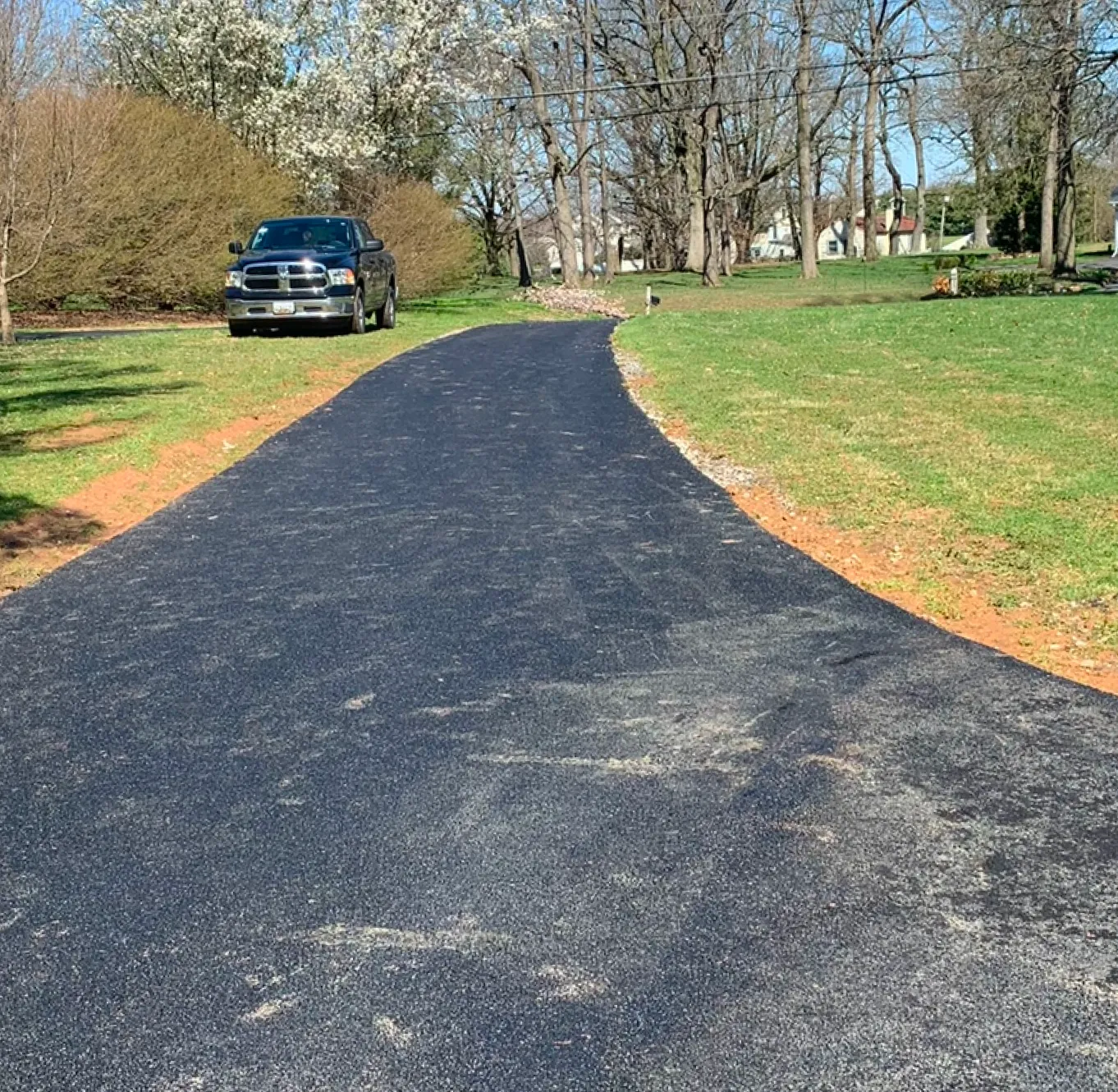A newly paved asphalt driveway curves toward a parked black pickup truck on a sunny day.