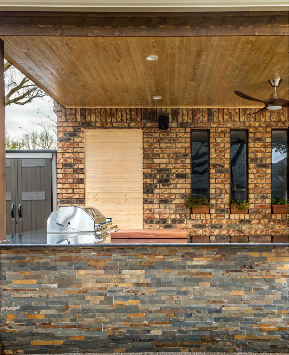 Outdoor kitchen with a grill, stone counter, brick wall, wood ceiling, and windows with potted plants.