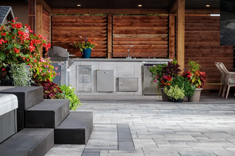 Outdoor kitchen with gray cabinets, grill, potted plants, and stone patio beside black steps.