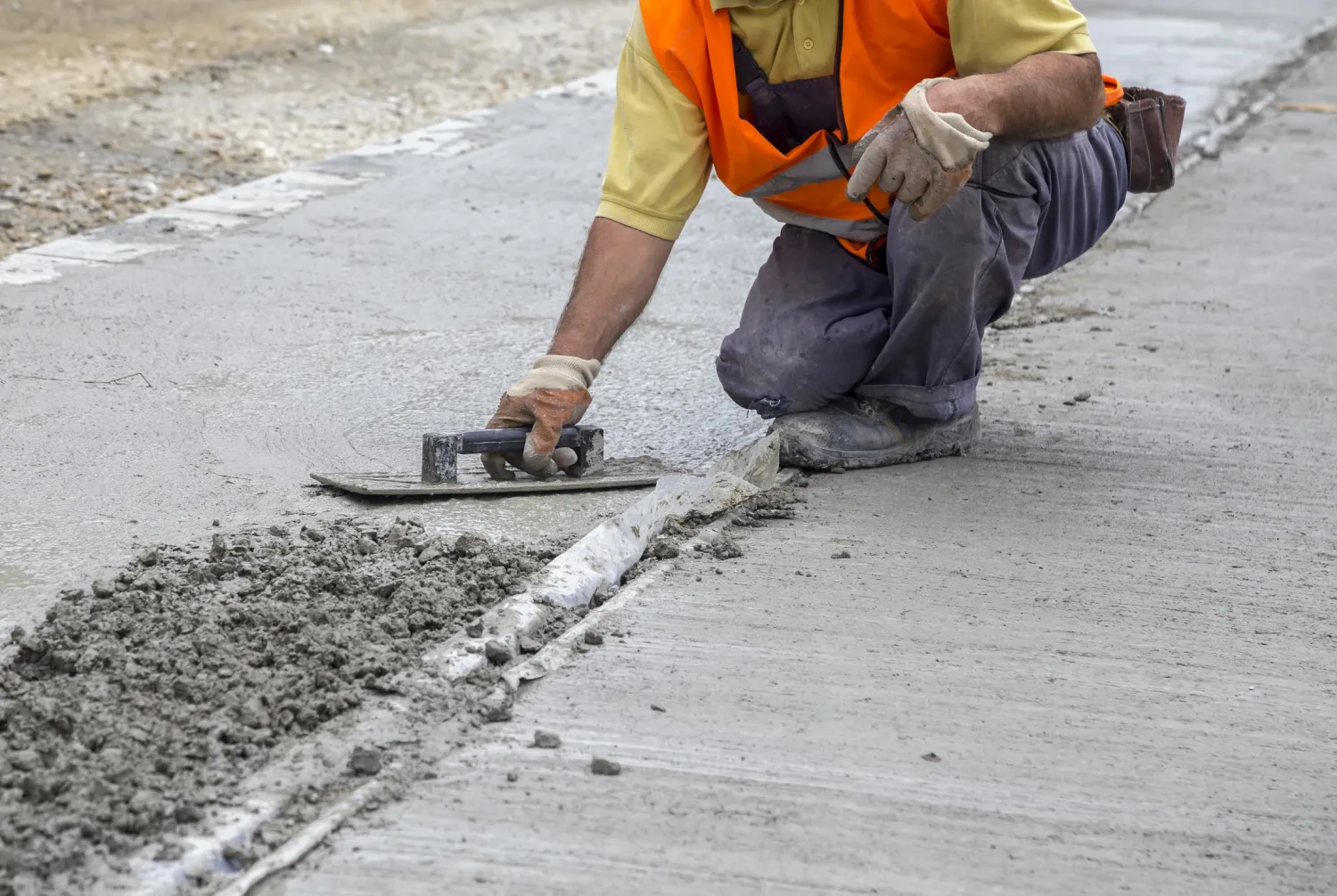 leveling-b Worker in orange vest spreading wet concrete on a road with a trowel, kneeling on one knee.