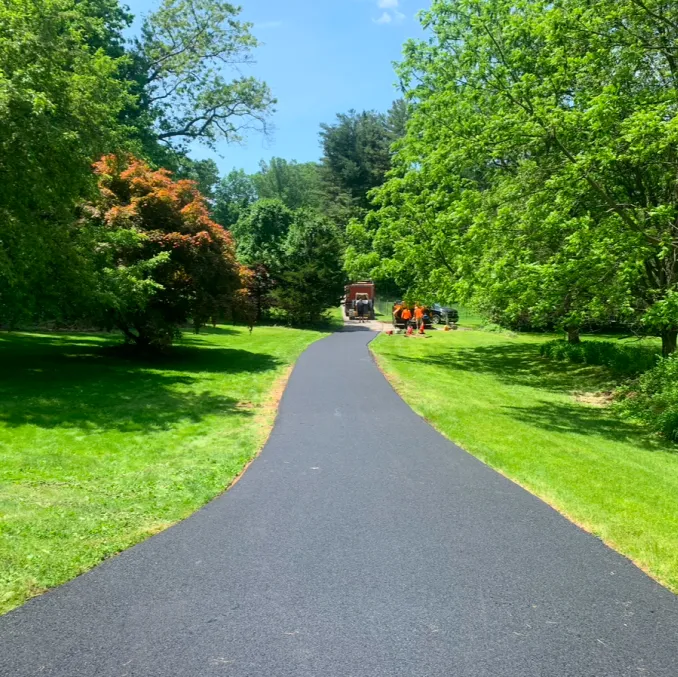 A newly paved black asphalt path curves through a green park with workers and vehicles ahead.