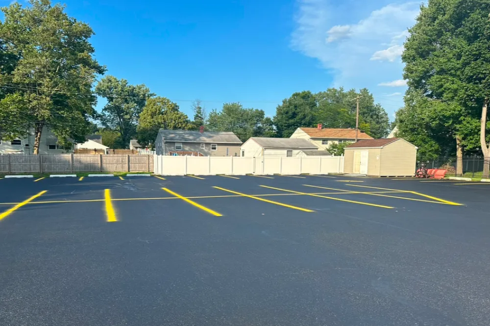 Freshly paved parking lot with bright yellow parking lines, surrounded by trees, houses, and a white fence.