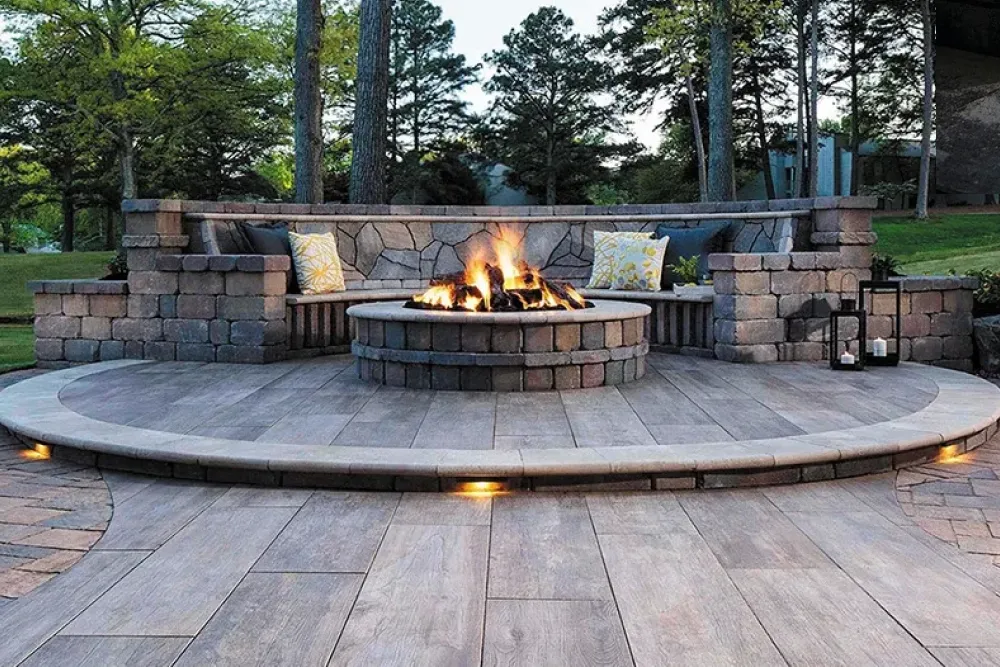 Stone fire pit with burning fire, curved stone bench, pillows, and trees in the background.