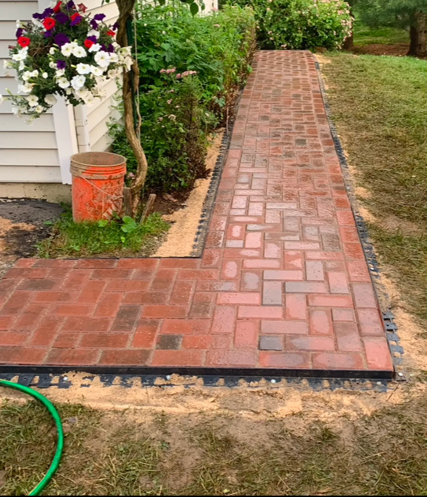 A newly laid brick path with sand edges beside a house, flowers, and a green garden hose.