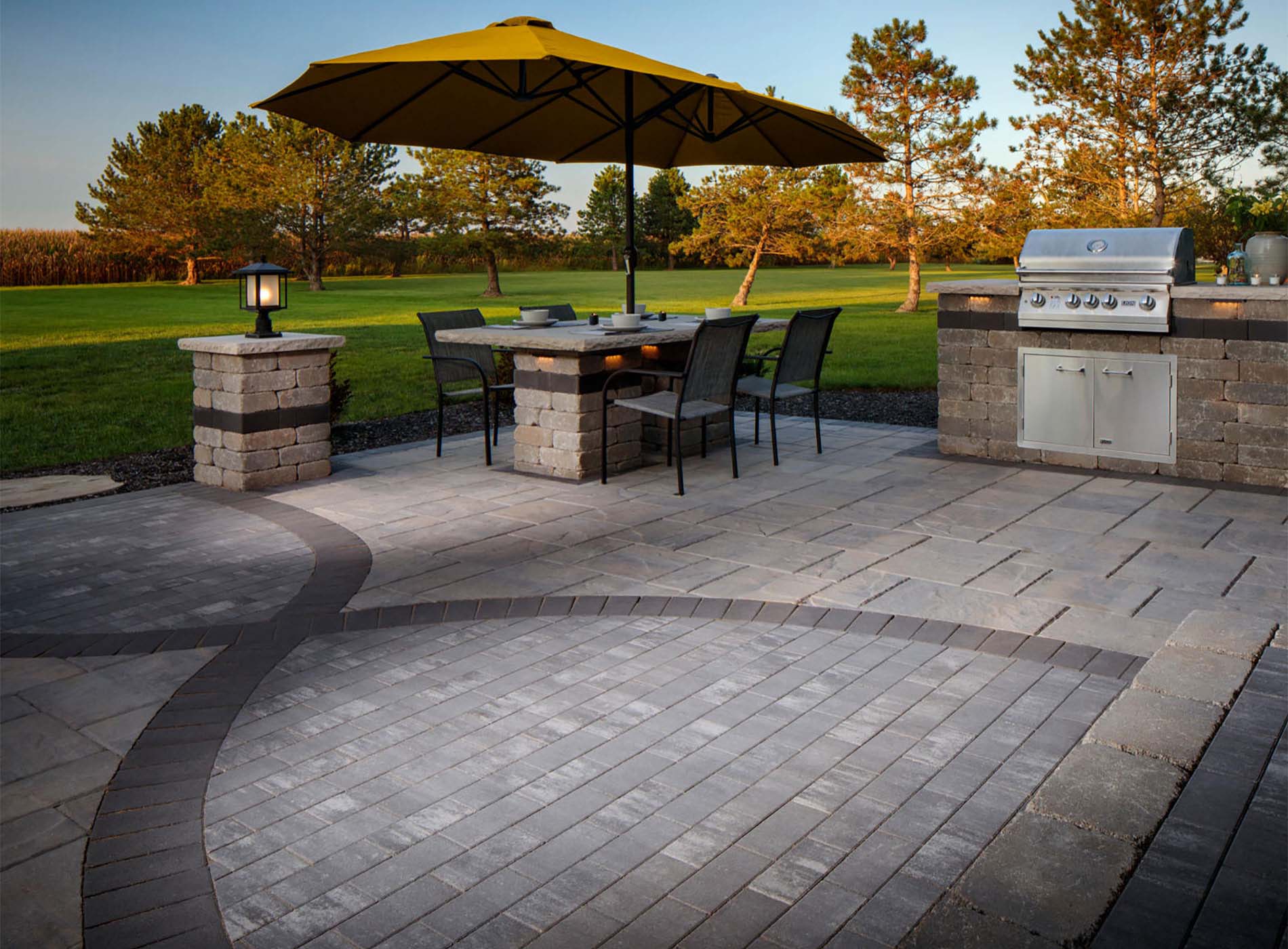 Patio with table, chairs, yellow umbrella, grill, lamp, and trees in the background on a sunny day.