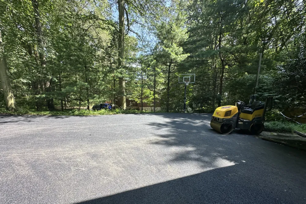 Freshly paved driveway with trees, a basketball hoop, and a yellow roller vehicle in bright sunlight.