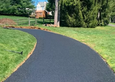 A freshly paved black asphalt path curves through a grassy yard with trees and a small shed.