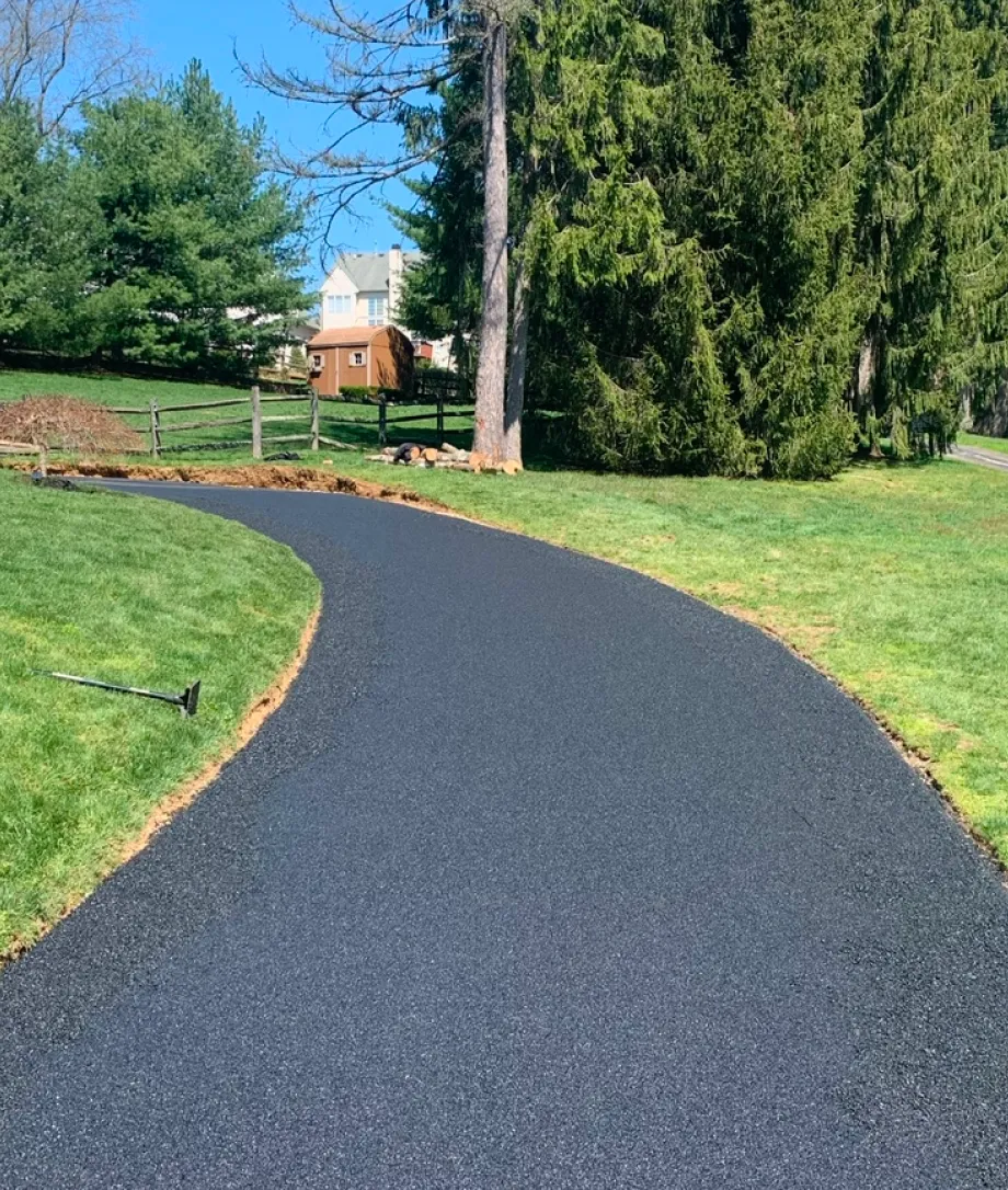 A freshly paved black asphalt path curves through a grassy yard with trees and a small shed.