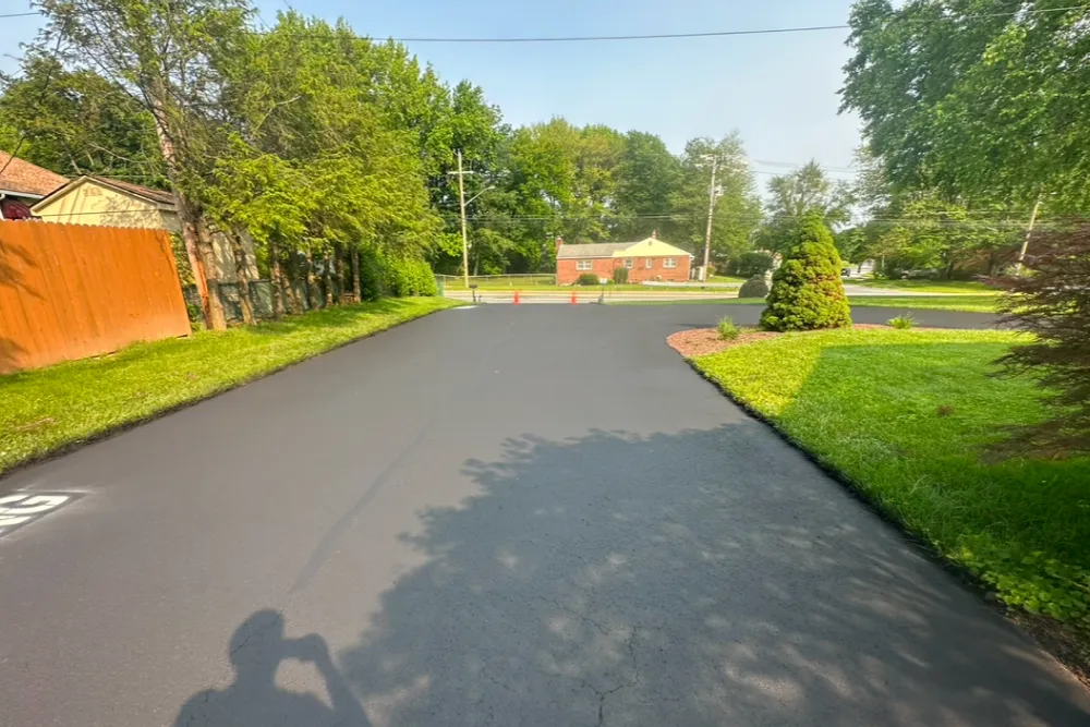 Freshly paved black asphalt driveway with green grass and trees, under a clear blue sky.