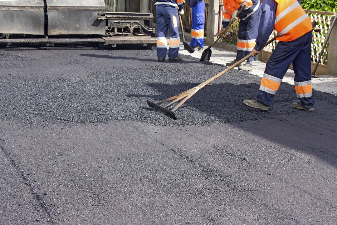 tristate-paving-infrared-asphalt-repair Workers in orange vests spread fresh asphalt on a road using rakes and machinery.