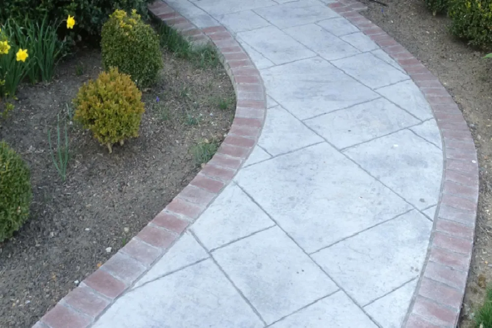 Curved concrete walkway bordered with red bricks, surrounded by dirt, shrubs, and blooming yellow flowers.