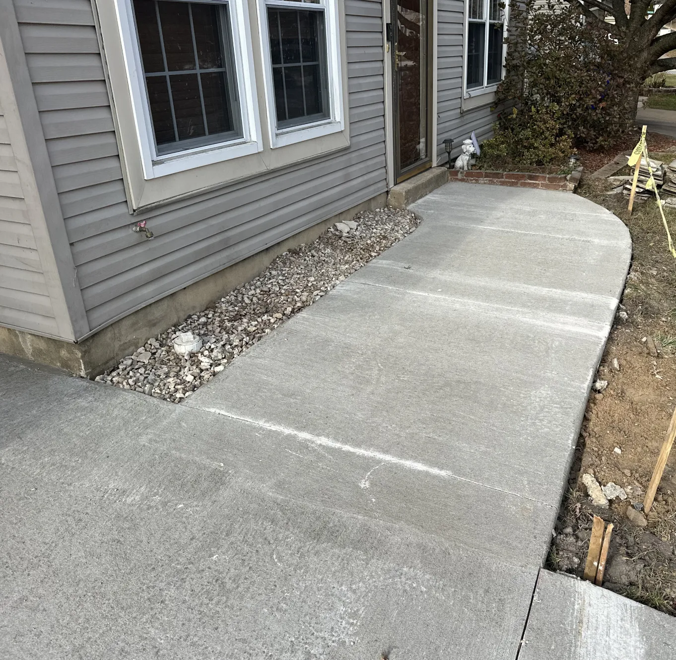 Freshly poured concrete sidewalk curves to a house entrance, with gravel along the house foundation.