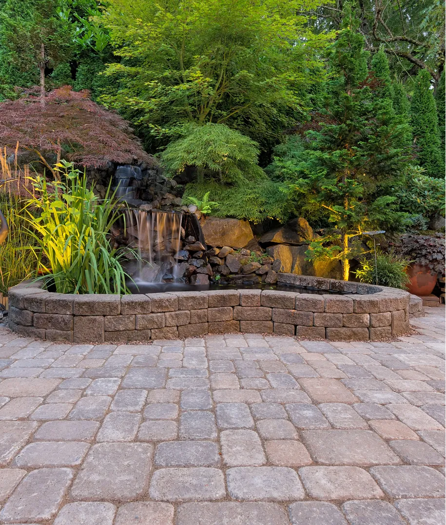 Stone patio with a raised planter, small waterfall, and lush green trees and plants in the background.