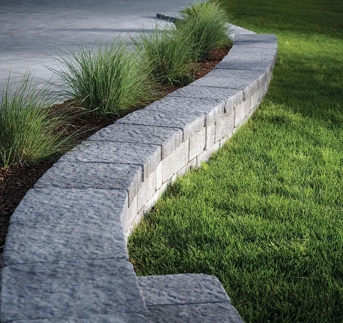 Curved stone retaining wall borders grass and concrete, with ornamental grasses planted along the wall.