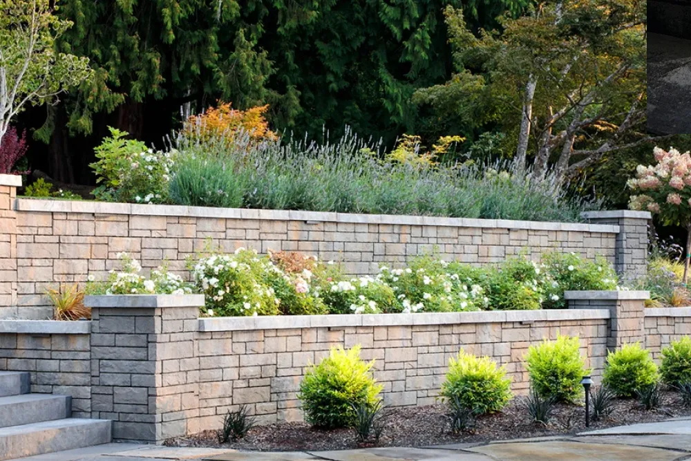 Tiered stone retaining walls with green shrubs, blooming white flowers, and trees in the background.