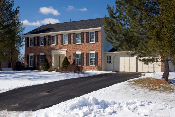 Tri-State-Paving-house-driveway-snow Two-story brick house with black shutters, attached garage, and driveway, surrounded by snow and trees.
