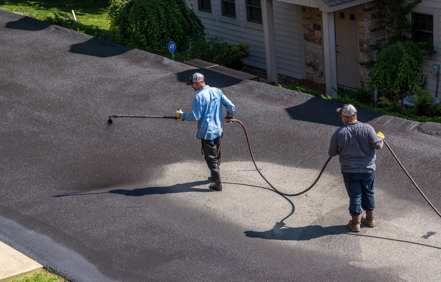 Two workers spray sealant on an asphalt driveway outside a house on a sunny day.